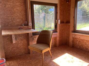 Sala de madeira bem iluminada com janelas grandes, uma mesa dobrável simples, dois pratos, um copo e uma cadeira xadrez verde. Aconchegante, minimalista, com luz natural e um ambiente tranquilo.
