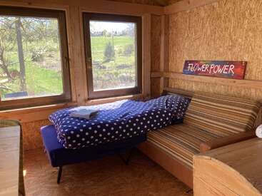 Um quarto aconchegante de madeira com janelas grandes, roupa de cama com bolinhas numa cama dobrável, sofá listrado, um letreiro com “Flower Power” e uma vista panorâmica verdejante lá fora.