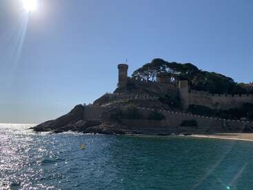 Una pintoresca fortaleza costera se alza sobre acantilados rocosos, rodeada de verdes árboles, con vistas a un mar azul centelleante. El sol brilla intensamente en el sereno cielo.