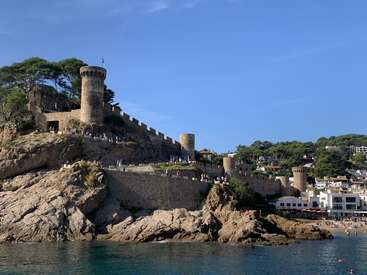 Un castillo medieval de piedra con varias torres redondas se asienta en lo alto de una colina rocosa, con vistas al mar azul y a una pequeña ciudad costera situada más abajo, bajo un cielo despejado.