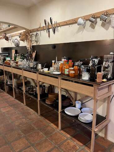 This cozy kitchen features open shelves with plates and cookware, a countertop filled with jars and bottles, and wall-mounted utensils, knives, and cups organized above.