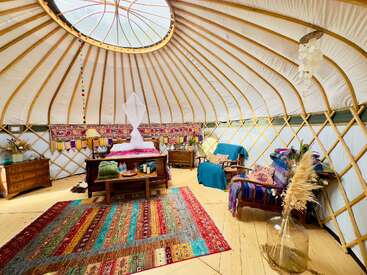 This cozy yurt features a colorful rug, comfortable bed, eclectic furniture, unique bohemian decor, natural light from a skylight, and inviting, warm wooden flooring throughout.