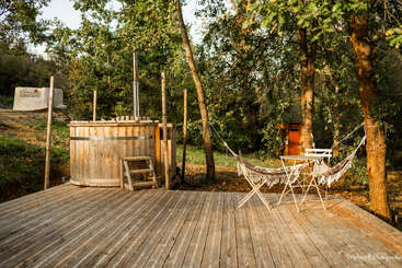 A peaceful wooden deck in a forest features a rustic hot tub, a small table with two chairs, and a cozy hammock strung between trees.