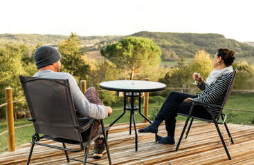 A man and woman relax on a wooden deck with scenic mountain views, sitting in chairs around a small table, enjoying the tranquil, natural surroundings together.