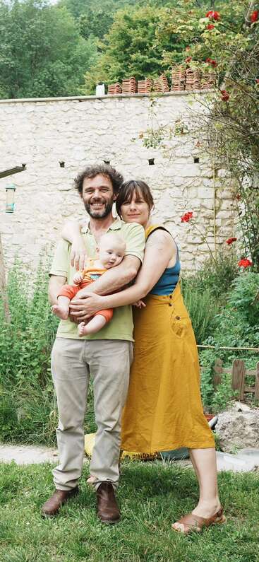 A happy family stands outdoors: father holding a baby, mother hugging both. Greenery, red flowers, and a rustic stone wall create a charming, peaceful background scene.