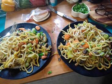 Das Bild zeigt zwei Teller mit Nudeln und Gemüse auf einem Holztisch, umgeben von Küchenutensilien und Zutaten. Die Teller sind schwarz und enthalten eine Vielzahl von Gemüsesorten.