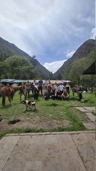 Un groupe de personnes pose en plein air dans une vallée pittoresque entourée de montagnes, en compagnie d'un cheval, d'un âne et d'un chien, sur une prairie, sous un ciel nuageux.
