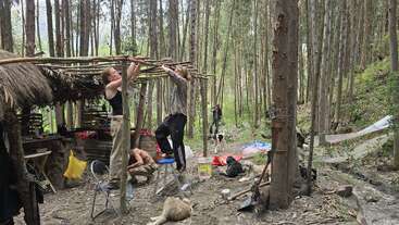 Deux personnes travaillent ensemble à la construction d'un abri au toit de chaume dans une forêt dense. Des chiens se reposent à proximité, tandis que des provisions et des outils sont éparpillés tout autour. D'autres personnes sont visibles plus loin à l'arrière-plan.