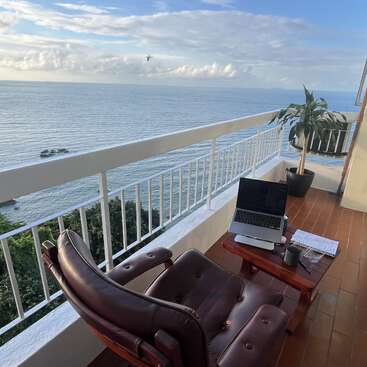 Um espaço de trabalho aconchegante na varanda tem uma cadeira marrom, uma mesa de madeira, um laptop, um notebook, um vaso de plantas e um café, com uma vista deslumbrante para o mar sob um céu claro.