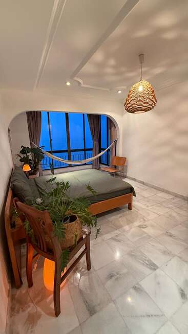 A cozy, modern bedroom with marble floors, large windows, green bedding, houseplants, wooden furniture, a hammock, woven pendant lamp, and a serene evening view.