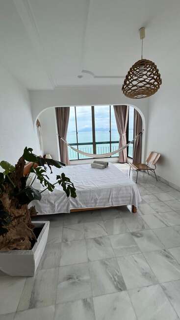 A bright, minimalist bedroom with marble floor, large plant, bed, hammock, and panoramic ocean view through floor-to-ceiling windows, creating a tranquil relaxing atmosphere.