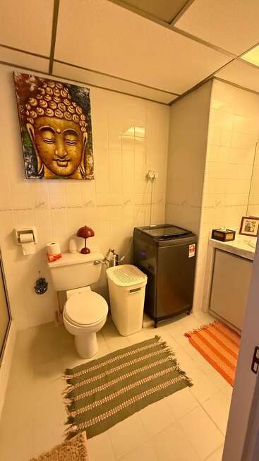 This bathroom features a toilet, washing machine, laundry basket, two rugs, a Buddha painting, a small lamp, and simple decor for a cozy atmosphere.