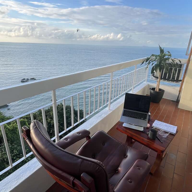 A cozy balcony workspace features a brown chair, wooden table, laptop, notebook, potted plant, and coffee, overlooking a stunning ocean view under a clear sky.