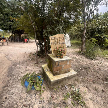 In einem bewaldeten Gebiet begrüßt ein Steinschild die Besucher der "Ferme Dessofa". Das Schild steht auf einem gemauerten Sockel, in dessen Nähe Pflanzen und eine rustikale Umgebung zu sehen sind.