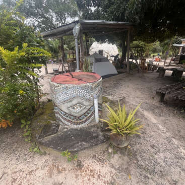 Eine rustikale Szene im Freien mit einem mosaikbedeckten Brunnen, Topfpflanzen, Holzbänken, einem Zelt, Bäumen und einem Holzunterstand, die eine friedliche, natürliche Umgebung schaffen.