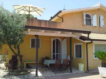 Une maison jaune à deux étages avec des fenêtres cintrées et un patio. Des meubles d'extérieur sont installés sous un toit. Un parasol et un arbre créent une atmosphère chaleureuse.