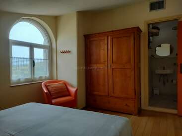 Une chambre confortable avec une grande fenêtre cintrée, un fauteuil orange, une armoire en bois et une salle de bains attenante. Les murs neutres créent une atmosphère chaleureuse, accueillante et simple.