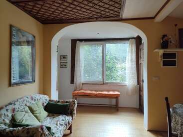 Uma sala de estar aconchegante com um sofá floral, almofadas verdes e um grande assento na janela. A luz natural preenche o espaço. Paredes amarelas quentes e detalhes em madeira por toda parte.