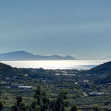 Eine ruhige Landschaft mit üppigen grünen Hügeln, einem glitzernden blauen Meer, in dem sich das Sonnenlicht spiegelt, fernen Bergen am Horizont und verstreuten Gebäuden im Tal.