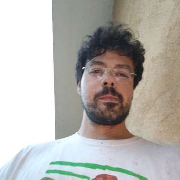 Un hombre con el pelo rizado y gafas lleva una camisa blanca. Tiene barba, mira directamente a la cámara y está de pie junto a una pared beige.