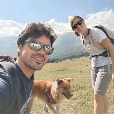 Un jeune homme et une jeune femme, portant tous deux des lunettes de soleil et un sac à dos, sourient en plein air avec leur chien par une journée ensoleillée, avec des montagnes et des champs herbeux derrière eux.