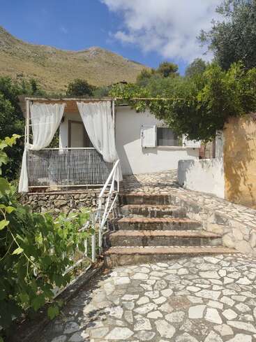 A quaint white cottage with a small stone patio and steps, surrounded by greenery, sits under a bright blue sky and hills in the background.