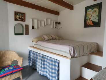 A cozy, elevated bed with striped bedding sits in a white room. There are wooden beams, a wicker chair, vibrant pillow, checkered curtain, and colorful artwork.
