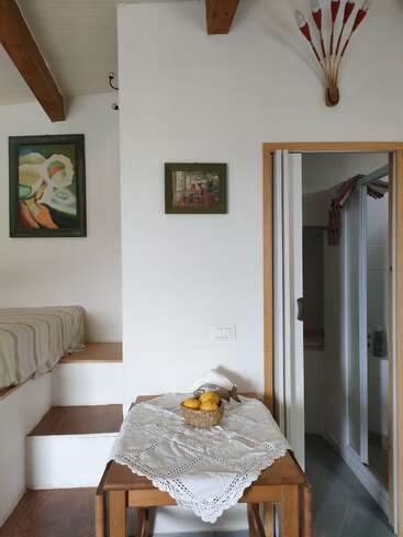 A cozy interior with exposed wooden beams, small bed, a wooden table covered by a lace cloth, basket of lemons, artwork, and an open doorway to a bathroom.