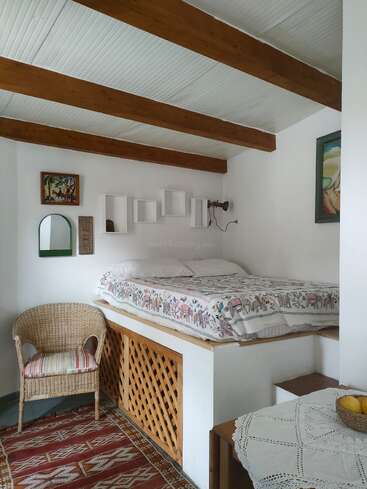 Um quarto aconchegante com uma cama elevada, uma cadeira de vime e um tapete colorido. A decoração inclui quadros nas paredes, prateleiras abertas e uma mesa com uma toalha de crochê.