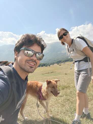 Un hombre y una mujer jóvenes, ambos con gafas de sol y mochila, sonríen al aire libre con su perro en un día soleado, con montañas y campos de hierba a sus espaldas.