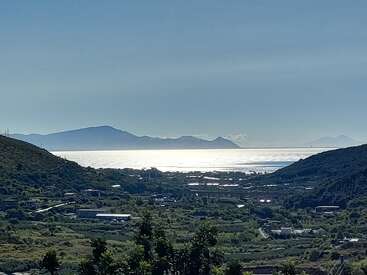 Uma paisagem serena com colinas verdejantes, um mar azul cintilante refletindo a luz do sol, montanhas distantes no horizonte e edifícios espalhados no vale abaixo.