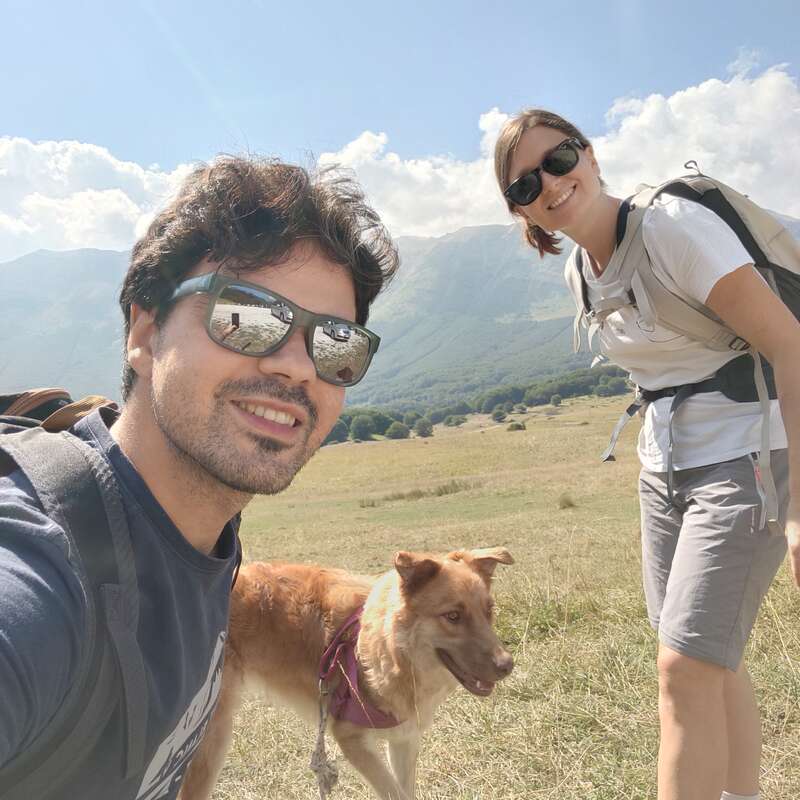 Um homem e uma mulher jovens, ambos usando óculos escuros e mochilas, sorriem ao ar livre com seu cachorro em um dia ensolarado, com montanhas e campos gramados atrás deles.