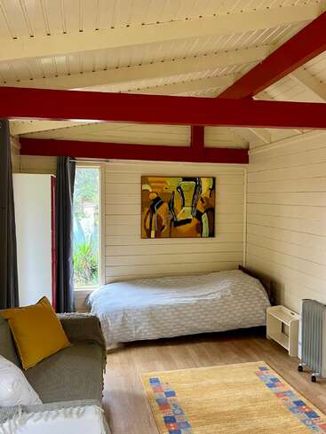 This cozy room features a single bed, sofa with pillows, colorful painting, light wood walls and ceiling, red beams, window, heater, and patterned rug.