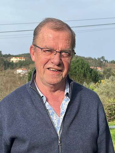 Ein Mann mittleren Alters mit Brille, der einen blauen Reißverschlusspullover über einem gemusterten Hemd trägt, steht an einem sonnigen Tag im Freien, hinter ihm sieht man Grünflächen und Hügel.