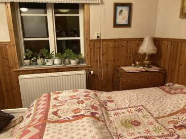 Une chambre chaleureuse avec des boiseries, un couvre-lit matelassé à motifs floraux, des plantes en pot sur le rebord de la fenêtre, une petite commode surmontée d'une lampe et des tableaux encadrés.