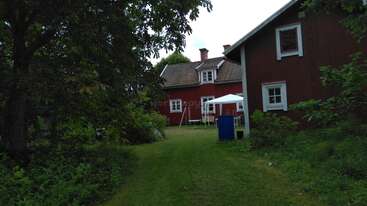 Une charmante maison en bois rouge nichée au milieu d'une végétation luxuriante et d'arbres, avec des moulures blanches, une pelouse, du mobilier d'extérieur et un chapiteau blanc. Un cadre paisible.