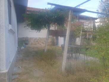 A imagem mostra um quintal rústico com grama crescida, uma pérgola, mesa e cadeiras ao ar livre, construções brancas, degraus de pedra e plantas verdes exuberantes, criando uma atmosfera aconchegante.