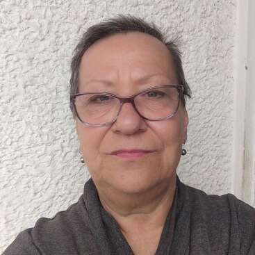 The image depicts an older woman with short dark hair, wearing glasses and a dark grey shirt, standing in front of a white wall with a textured surface.