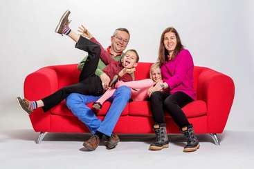 L'image montre une famille de quatre personnes posant pour une photo, avec deux adultes et deux enfants, tous souriants et regardant l'appareil, devant un fond uni.