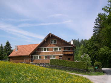 L'image représente une grande maison à deux étages, à l'extérieur marron et au toit orange, située dans un champ herbeux serein entouré d'arbres et d'un ciel bleu.