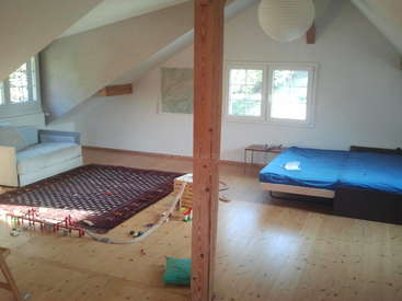L'image représente une pièce spacieuse au plafond incliné, avec un lit bleu, un canapé blanc, un tapis et des jouets éparpillés sur le sol en bois.