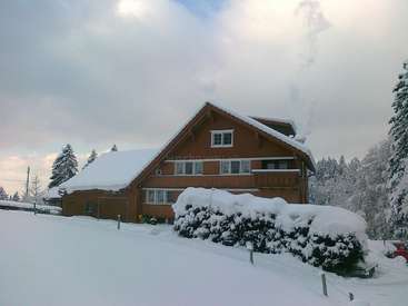 L'image représente une grande maison à deux étages avec un toit à pignon, couverte de neige, entourée d'arbres et d'un ciel nuageux, dégageant une atmosphère hivernale sereine.