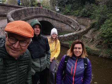 Quatro pessoas vestidas calorosamente posam em um canal, em frente a uma ponte de pedra curva cercada por vegetação. Eles parecem felizes, curtindo uma aventura ao ar livre juntos.