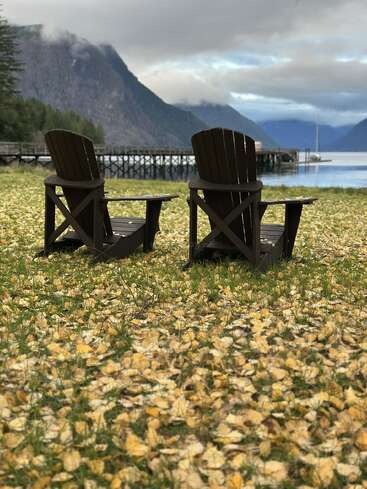 Two Adirondack chairs sit on leaf-covered grass, facing a tranquil lake. Mountains and a dock stretch into the distance under a dramatic, cloudy sky. Peaceful atmosphere.