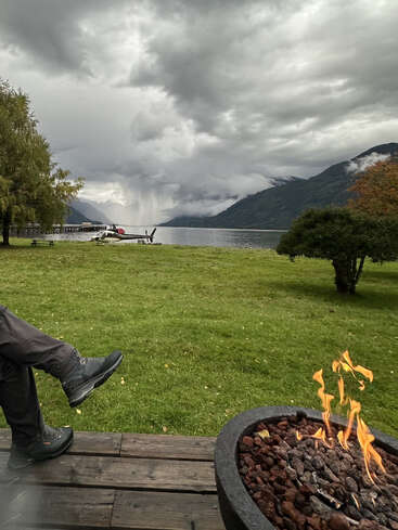 A cozy lakeside scene with a person’s boots resting on a wooden deck, fire pit ablaze, cloudy skies, mountains, green grass, and a docked seaplane.