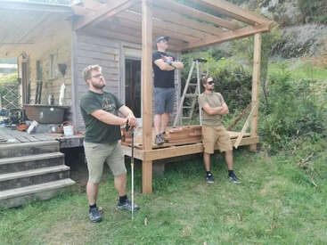Three men stand proudly by a partially constructed wooden porch beside a rustic house, surrounded by tools, building materials, greenery, and a ladder in the background.