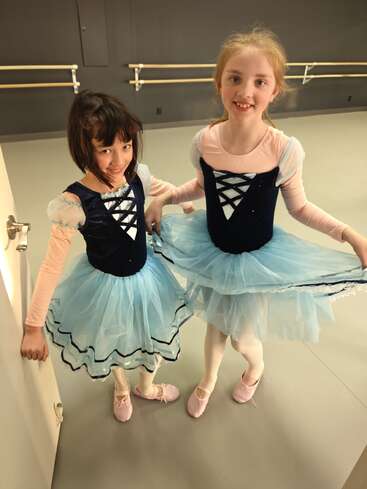 Dos niñas con tutús azules de ballet y medias rosas sonríen en un estudio de danza, listas para la clase de ballet, de pie juntas sobre el suelo liso y gris.