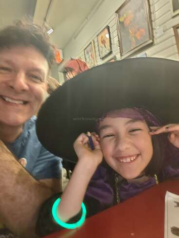 Un hombre y un niño sonríen felices para hacerse un selfi. El niño lleva un gran sombrero negro, un traje morado y una pulsera verde brillante. Detrás de ellos hay cuadros colgados.