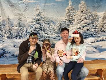 Cuatro personas están sentadas en un banco, sosteniendo accesorios divertidos ante un fondo de bosque nevado en invierno, sonriendo y disfrutando juntas de una foto festiva y alegre.