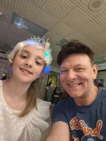 Un hombre sonriente y una niña con un vestido blanco y una tiara se hacen juntos una alegre selfie en el interior, con luces y adornos festivos al fondo.